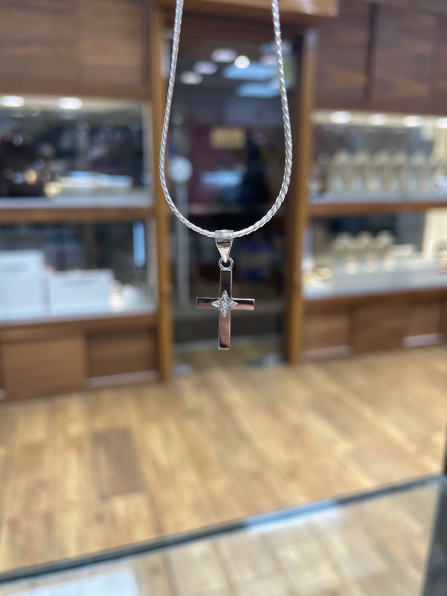 A silver cross pendant necklace hanging from a silver chain, set against a blurred jewelry store background with wooden display cases and a wood floor.